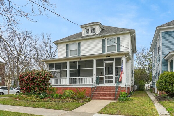 A two-story house with a front porch, a brick foundation, and a small garden with flowering plants.