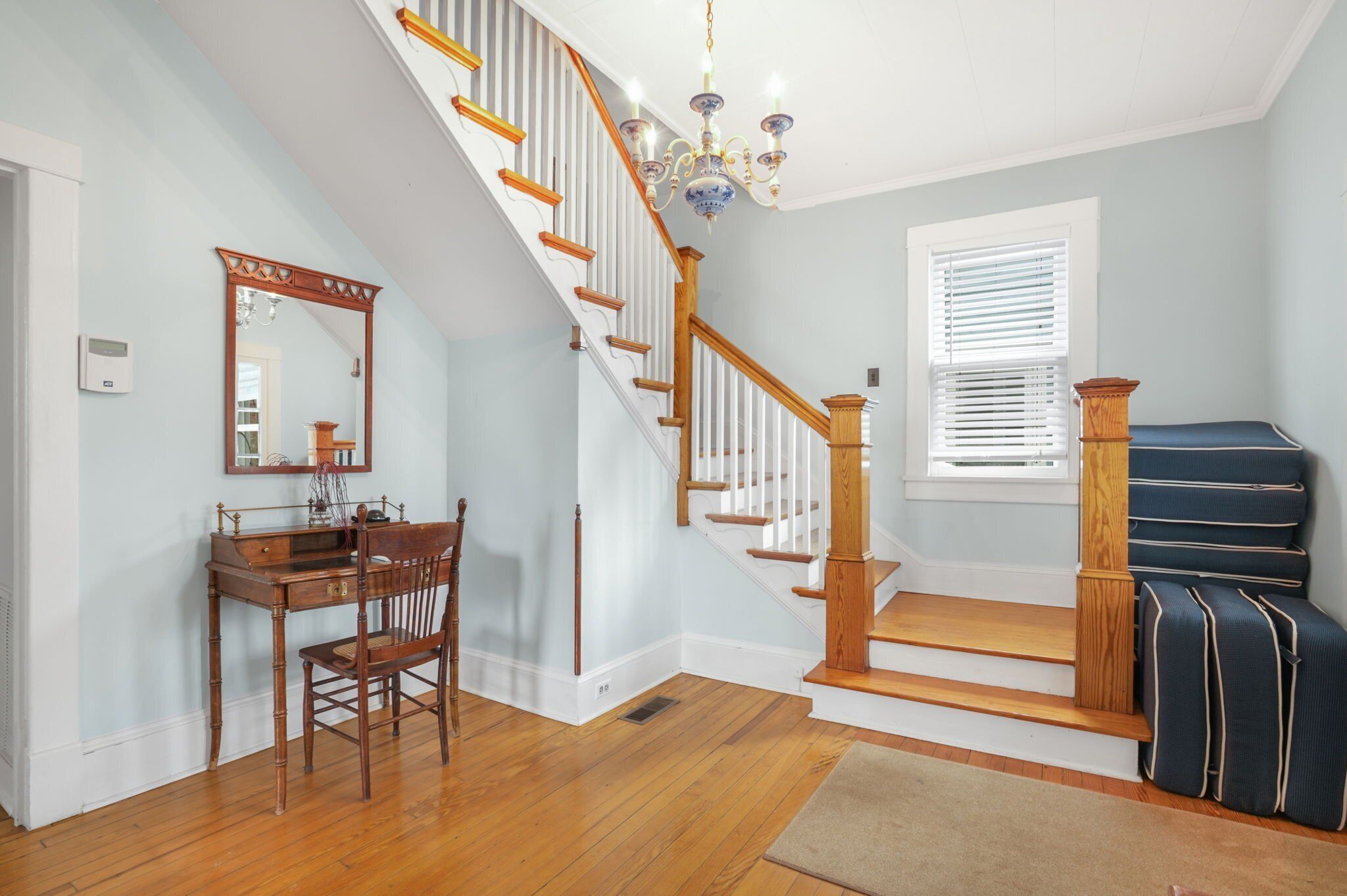 A staircase with wooden railings and a chandelier, a desk with a chair, and a window with blinds.