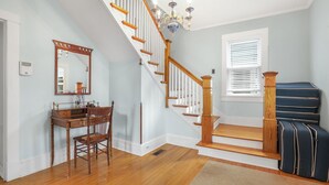 Un escalier avec des rampes en bois et un lustre, un bureau avec une chaise, et une fenêtre avec des stores.