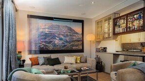 Ein Wohnzimmer mit großem Fernseher, einer Couch, zwei Sesseln, einem Couchtisch und einer Küchenzeile mit Schränken und einer Arbeitsplatte.