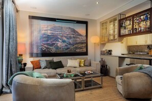 A living room with a large TV, a sofa, two armchairs, a coffee table, and a kitchen area with cabinets and a countertop.