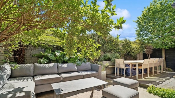 Eine Terrasse mit einer Couch, Tisch und Stühlen, umgeben von Pflanzen und einer Backsteinmauer.