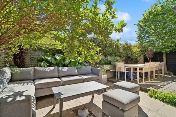 A patio with a sofa, table, and chairs, surrounded by greenery and a brick wall.