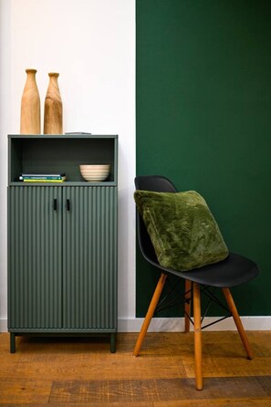 A green cabinet with two wooden vases, a bowl, and books. A green chair with a cushion is next to it.