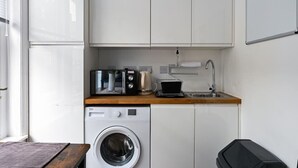 A compact kitchen with a washing machine, a coffee maker, and a microwave on the countertop. - Central London | King Bed | Smart Tv | Big Windows (London)