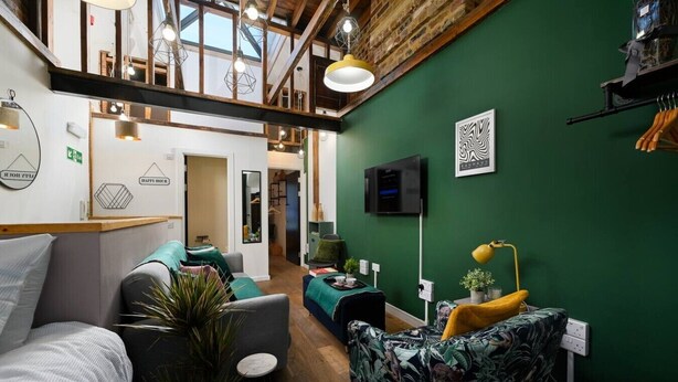 A modern interior with a green wall, wooden beams, a sofa, a chair, and a TV mounted on the wall. - Central London | King Bed | Smart Tv | Big Windows (London)
