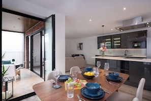 Ein moderner Essbereich mit einem Holztisch, gedeckt für vier Personen, eine Küche mit Edelstahlgeräten und eine Schiebeglasdoor zu einem Balkon.