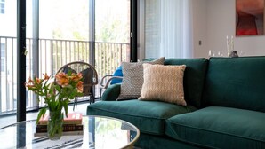 Un canapé vert agrémenté de coussins à motifs, une table basse en verre sur laquelle est posé un vase de fleurs, et un fauteuil offrant une vue sur l’extérieur.