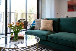 Un sofá verde con almohadones estampados, una mesa de centro de vidrio con un jarrón de flores, y una silla con vistas al exterior.