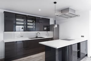 Una cocina moderna con armarios oscuros, una encimera blanca, un refrigerador de acero inoxidable y una lámpara colgante.