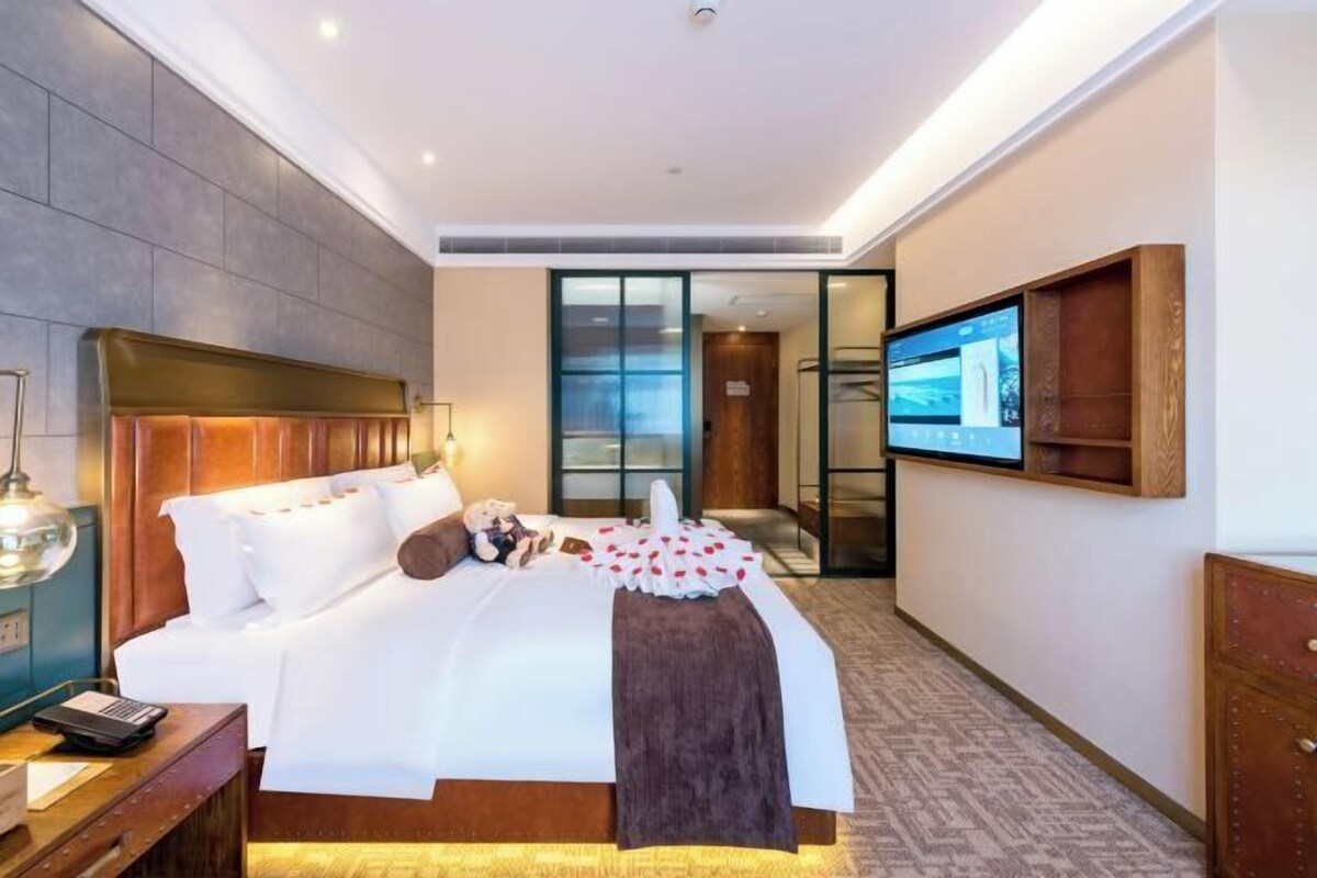 A hotel room with a bed, bedside table, lamp, TV, and a view of the outside.