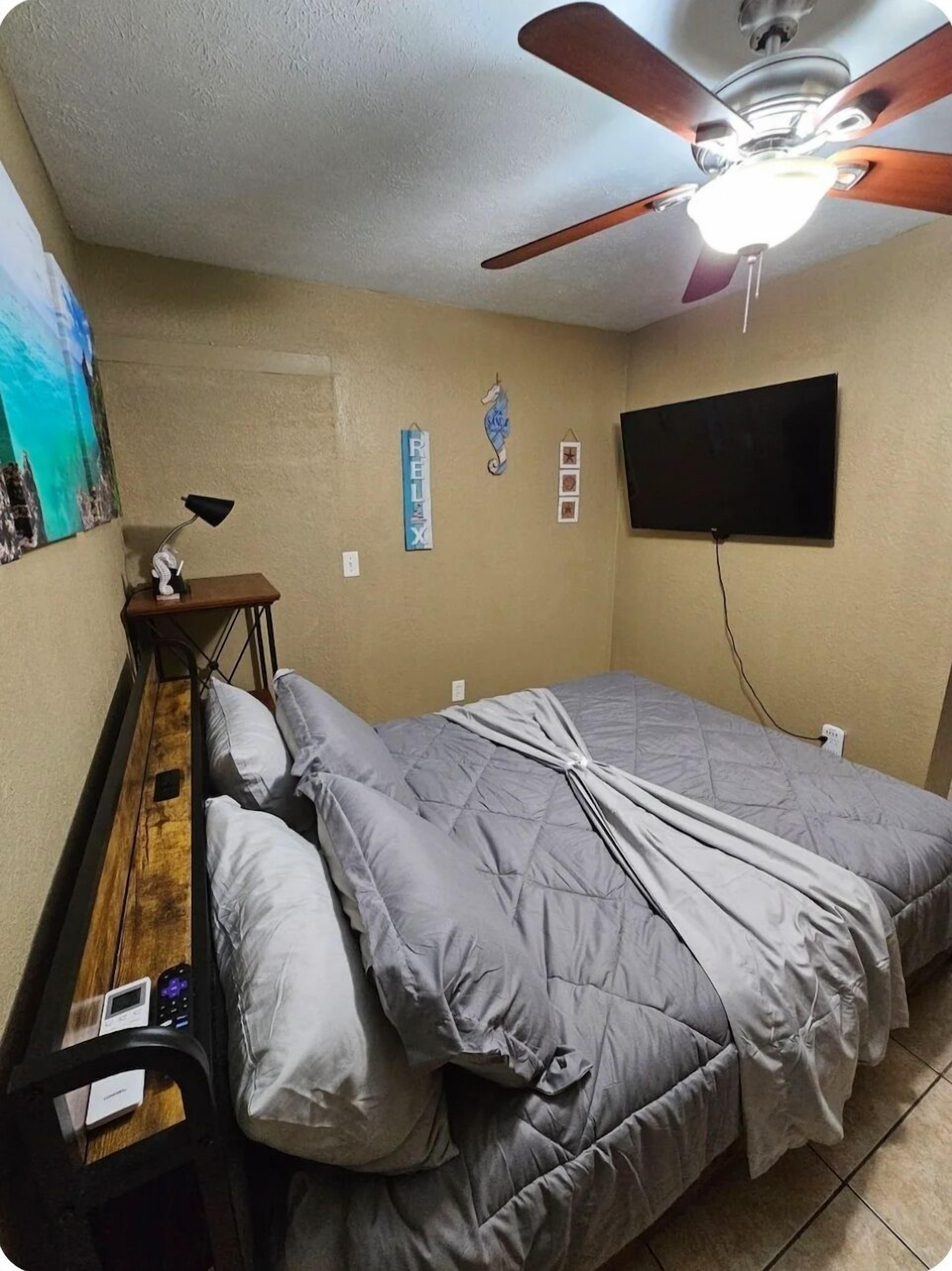 A bedroom with a bed, a desk, a chair, a ceiling fan, a TV, and a wall with a painting and decorations.