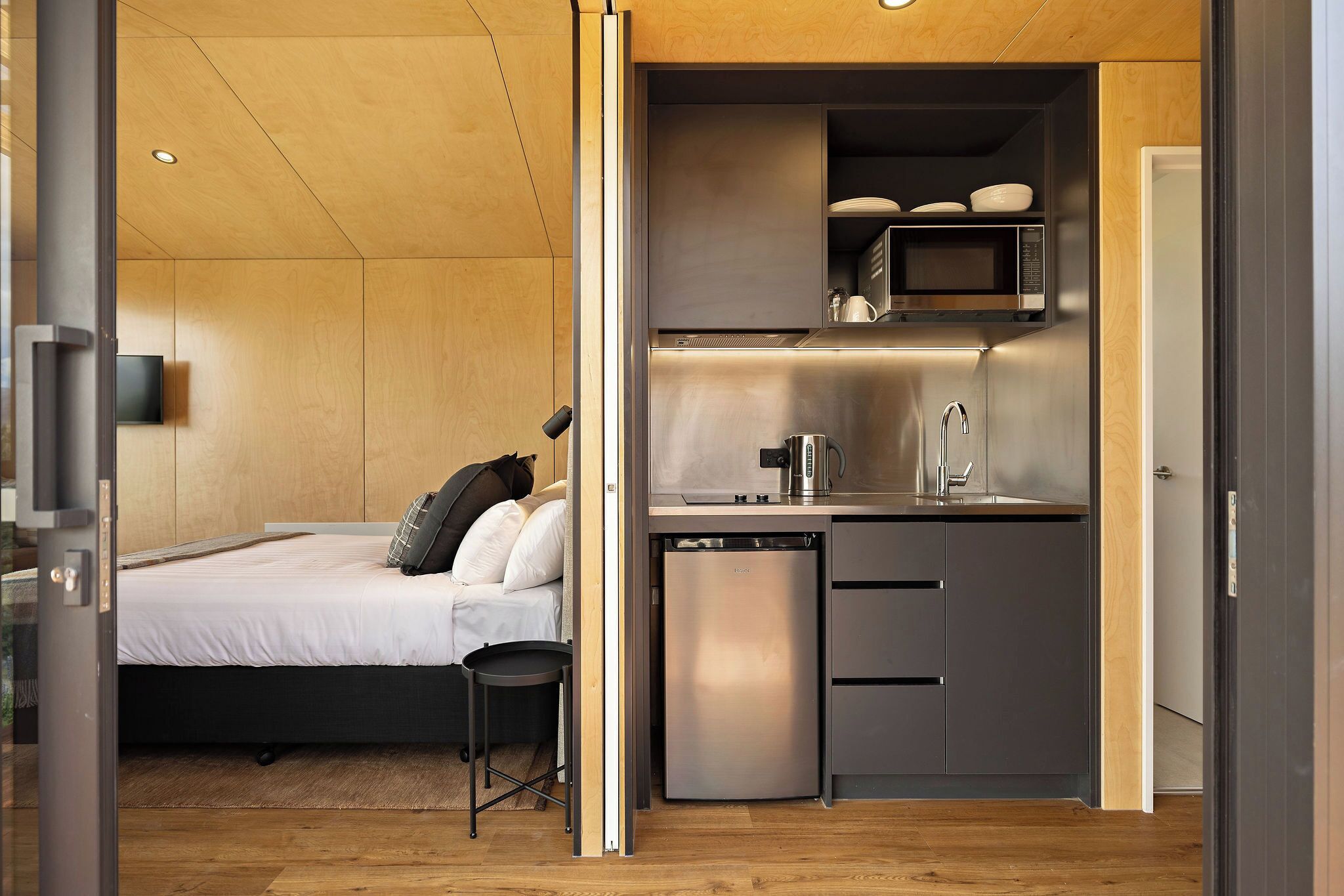Ein kleines Schlafzimmer mit Bett, einem kleinen Hocker und einer integrierten Küchenzeile inklusive Mikrowelle, Spüle und Kühlschrank.