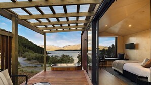 Une chambre moderne dotée d’une grande fenêtre, d’un lit, d’une télévision et d’un jacuzzi sur une terrasse en bois offrant une vue sur un lac et des montagnes.