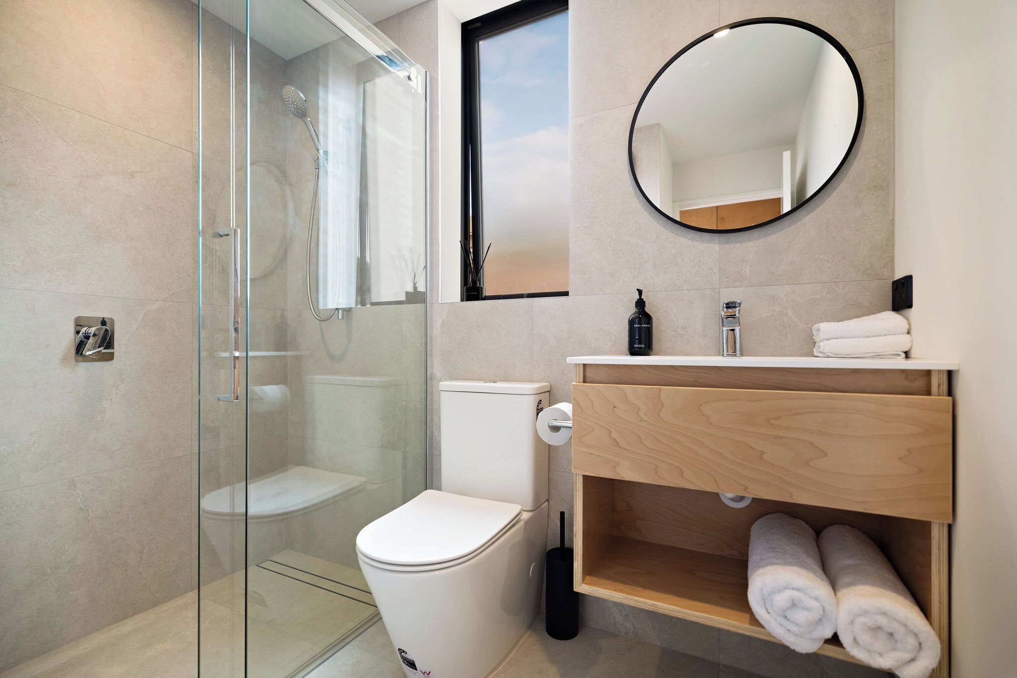 A modern bathroom with a glass-enclosed shower, a white toilet, a wooden vanity with a mirror, and neatly rolled towels.