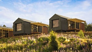 Deux chalets modernes dotés de grandes fenêtres et de terrasses en bois, nichés au milieu d’un champ de fleurs violettes.
