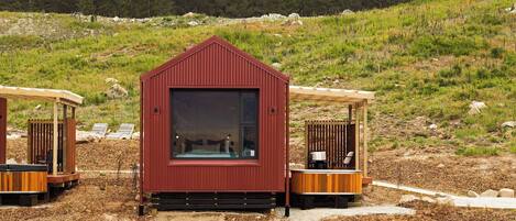 A red cabin with a large window, surrounded by a wooden deck and a forested hillside.