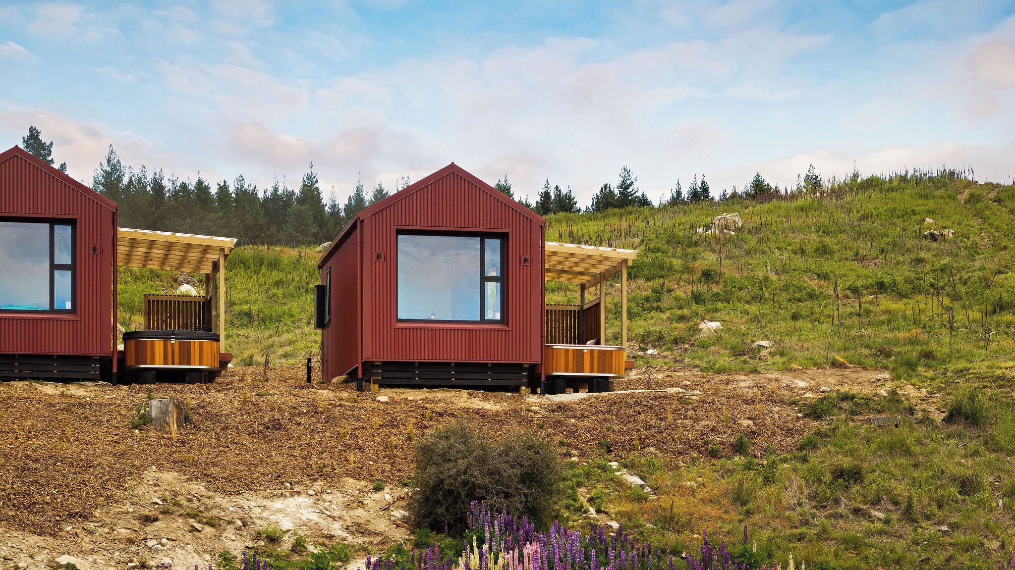 Two red cabins with large windows and wooden decks, situated on a grassy hillside with a clear sky.