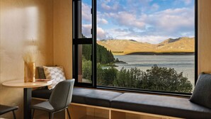 Une pièce dotée d’une grande fenêtre donnant sur un lac et des montagnes, une table ronde avec une chaise, et un banc avec des coussins.