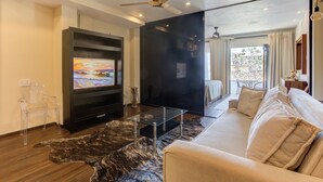 A modern living room with a flat-screen TV, a glass coffee table, a leather rug, a sofa, and a ceiling fan.