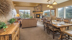A cozy living room with a fireplace, a dining area with wooden furniture, and large windows offering a view of the outdoors.