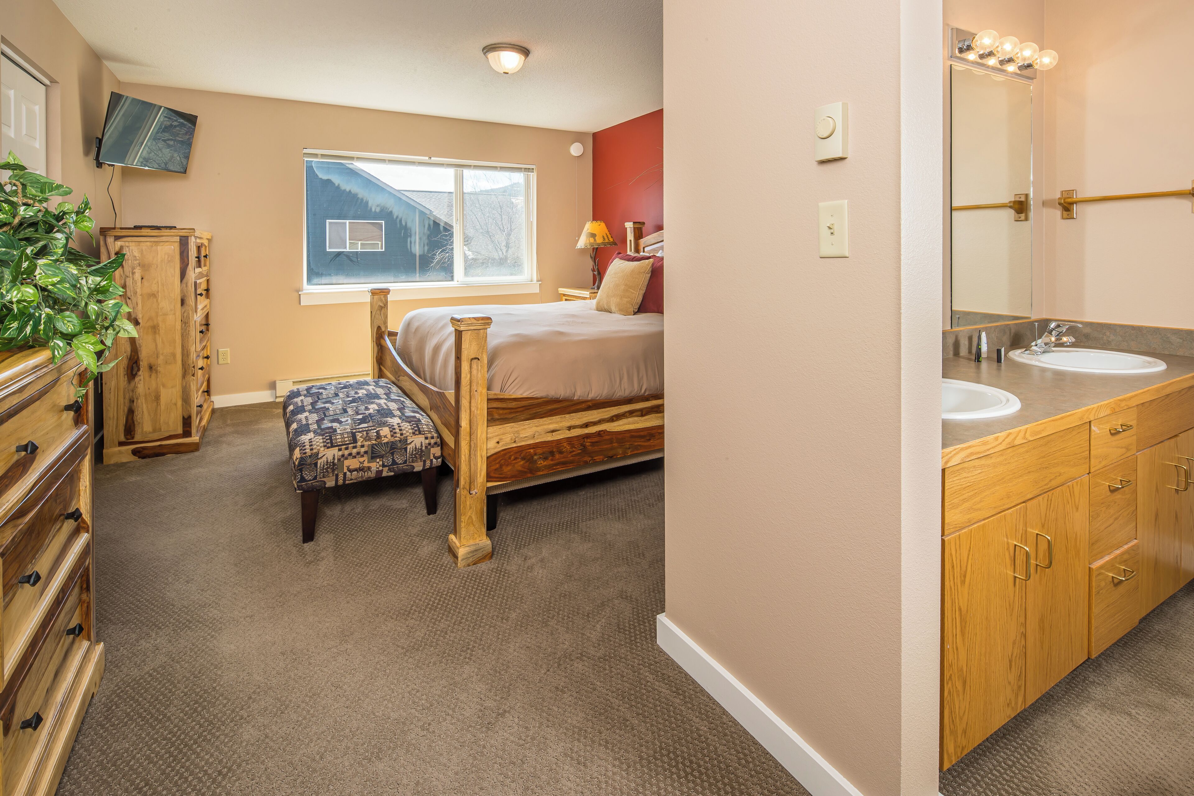 Une chambre à coucher comprenant un lit, une commode en bois, un banc, une fenêtre et une salle de bain avec un lavabo et un miroir.