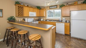A kitchen with wooden cabinets, a central island with stools, a microwave, a refrigerator, and a window with a view of a house.