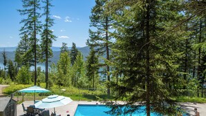 Zona de piscina con vista a un lago y montañas, rodeada de árboles altos.