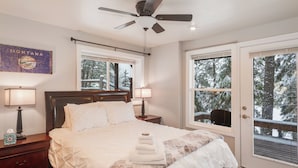 Un dormitorio con cama, mesitas de noche, ventilador de techo y vistas a árboles nevados a través de las ventanas.