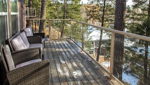 Terraza de madera con mobiliario de mimbre y barandilla de vidrio, con vista a un área boscosa.
