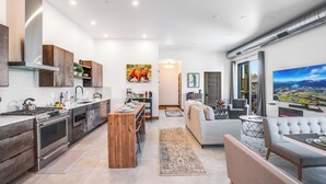 A modern kitchen with stainless steel appliances, a wooden island, and a dining area with a long table. There is a living room with a gray sofa and a large TV. - Located in Downtown Whitefish,Walk to Dining and Shops- Nearby Lake and Ski! (Whitefish)