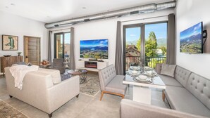 A modern living room with a sofa, a dining table set for four, and a large window with a scenic view. - Located in Downtown Whitefish,Walk to Dining and Shops- Nearby Lake and Ski! (Whitefish)
