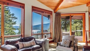 Un vaste espace de vie comprenant un canapé en cuir, un fauteuil gris et une table basse en verre. De grandes fenêtres offrent une vue sur une terrasse et sur le paysage extérieur.