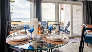 A dining area with a glass table, plates, and blue wine glasses, overlooking a beach.