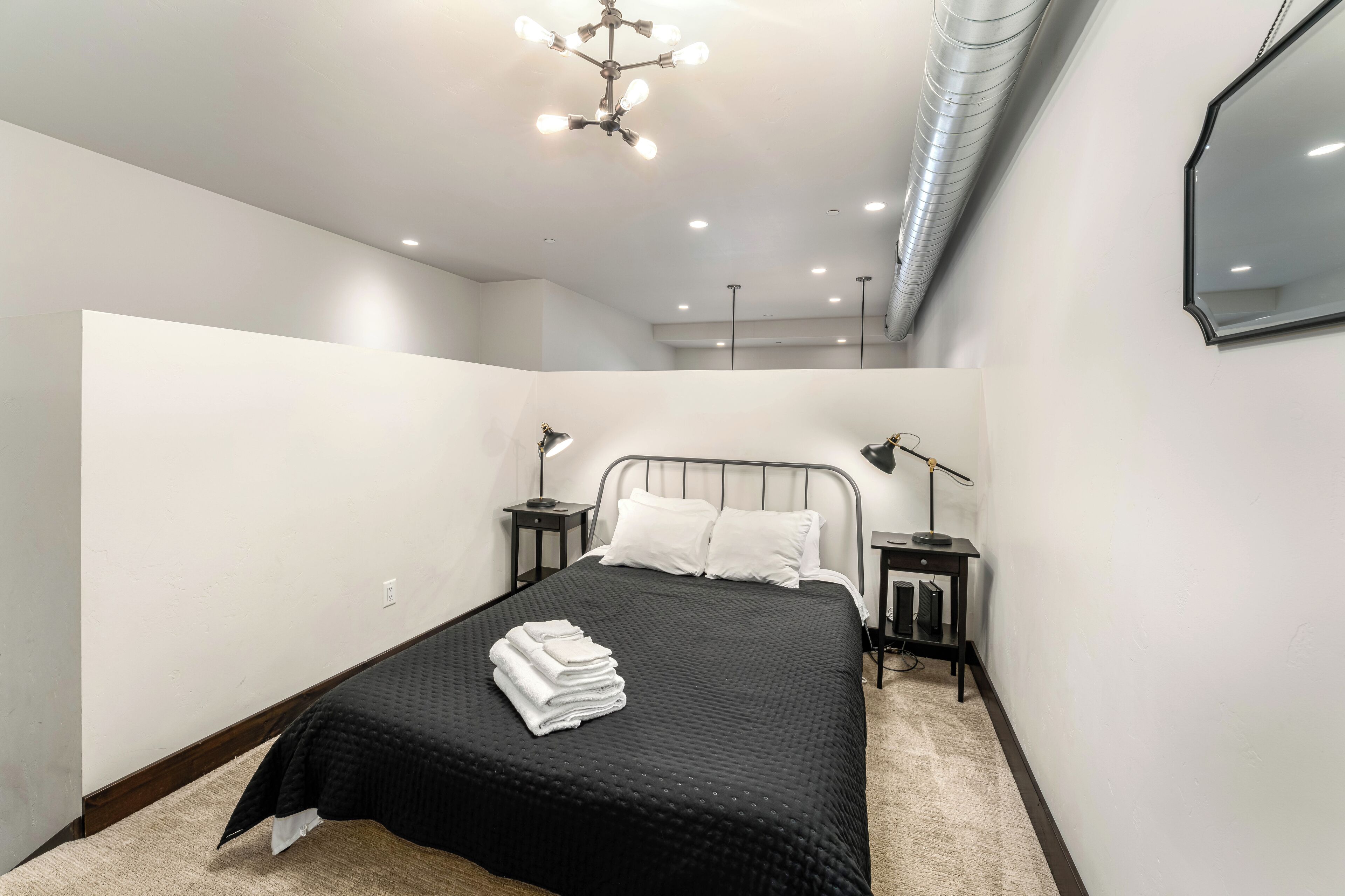Un dormitorio moderno con una colcha negra, almohadas blancas, una mesita de noche negra con una lámpara y una lámpara de techo.