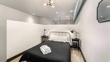 Un dormitorio moderno con una colcha negra, almohadas blancas, una mesita de noche negra con una lámpara y una lámpara de techo.