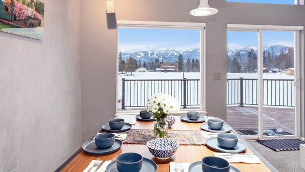 Un comedor con una mesa preparada para seis comensales, ventanas amplias con vista a la montaña y un cuadro en la pared.