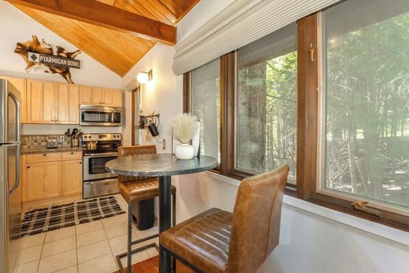 Una cocina con armarios de madera, electrodomésticos de acero inoxidable, una mesa de comedor con sillas y vistas al bosque a través de un amplio ventanal.