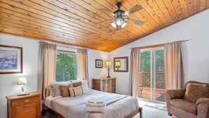 Ein Schlafzimmer mit Holzbalkendecke, einem Bett mit beiger Bettdecke, einem Nachttisch, einer Kommode und Blick ins Freie durch eine Schiebetür.