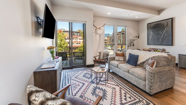A spacious living room with a large sofa, a coffee table, and a TV mounted on the wall. There is a large window with a view of buildings outside. - Located in Heart Downtown Whitefish, Walking Distance to River + Short Drive to Skiing! (Whitefish)