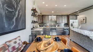 A modern kitchen with dark wood cabinets, stainless steel appliances, and a dining area with a wooden table set for two. - Located in Heart Downtown Whitefish, Walking Distance to River + Short Drive to Skiing! (Whitefish)