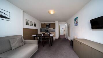 Une chambre d’hôtel compacte comprenant un canapé, une table à manger avec des chaises, un coin cuisine, un téléviseur fixé au mur et un petit meuble de rangement.
