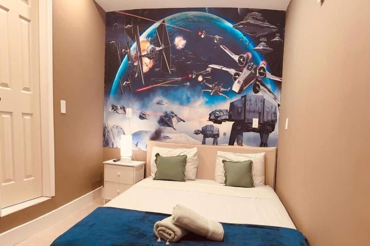 Quarto com cama, almofadas, mesa de cabeceira e um mural na parede com tema espacial.