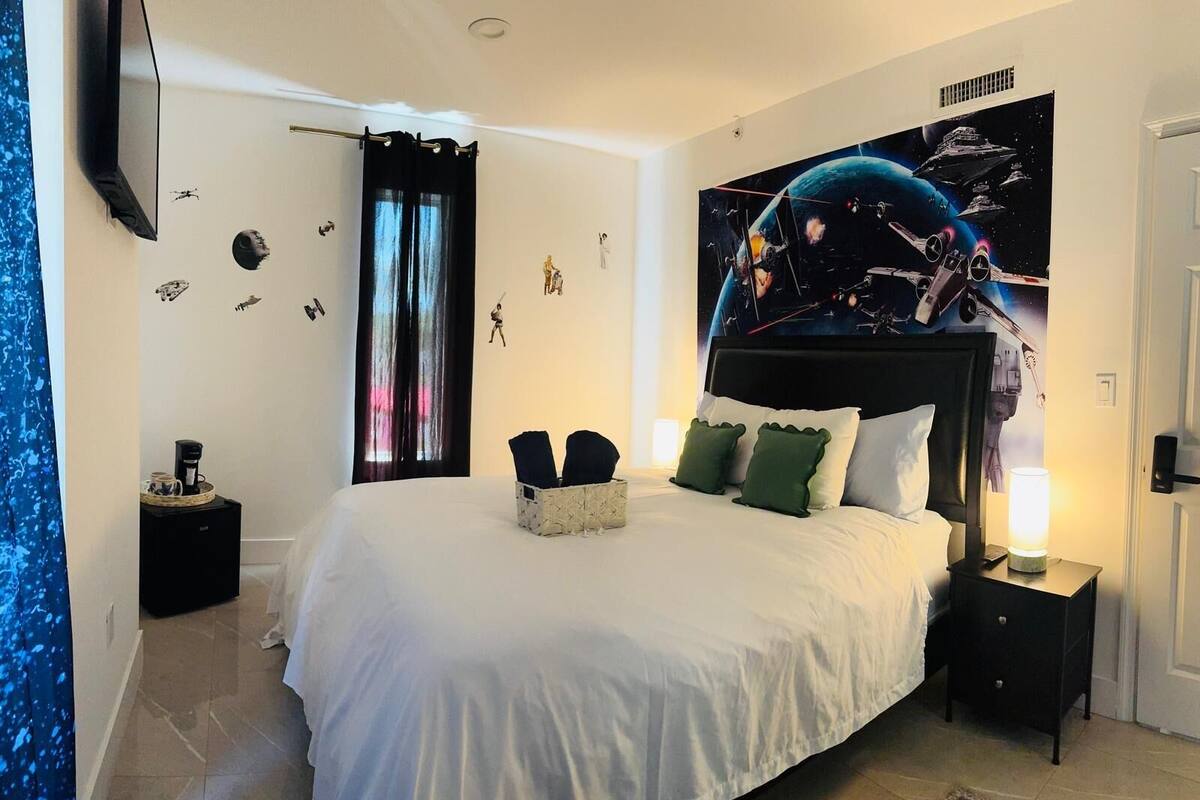 Quarto com uma cama grande, uma mesinha de cabeceira com luminária, parede decorada com tema espacial e janela com cortinas.