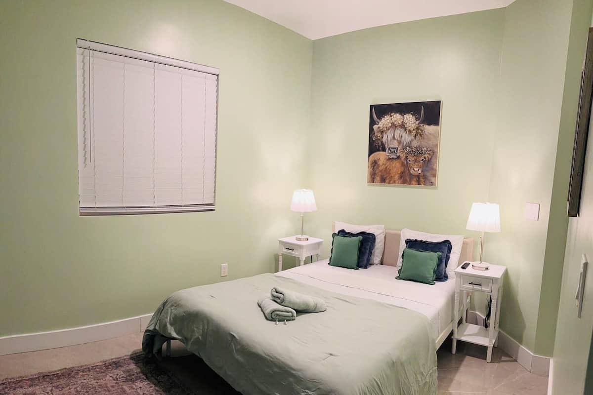 Quarto com cama, duas mesas de cabeceira, um criado-mudo, janela com persianas e um quadro na parede.