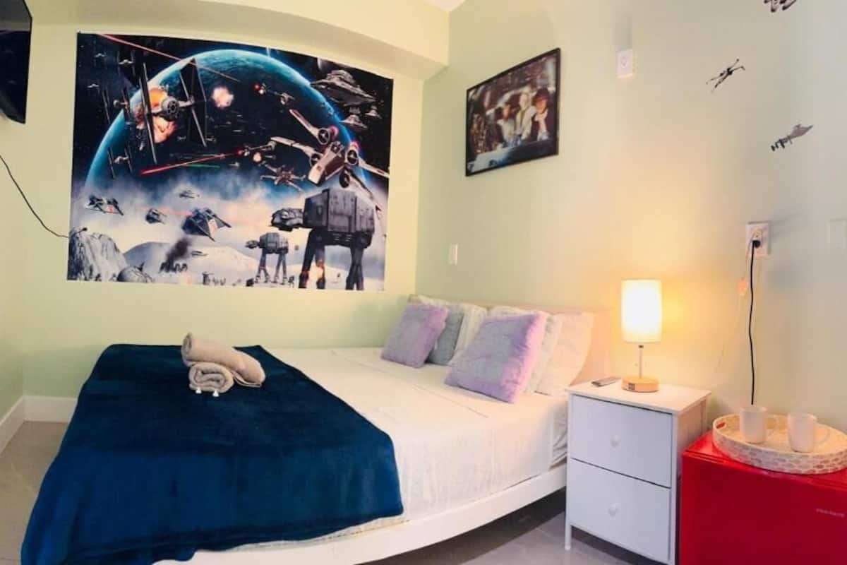 Quarto com cama, mesa de cabeceira, luminária e um mural na parede.