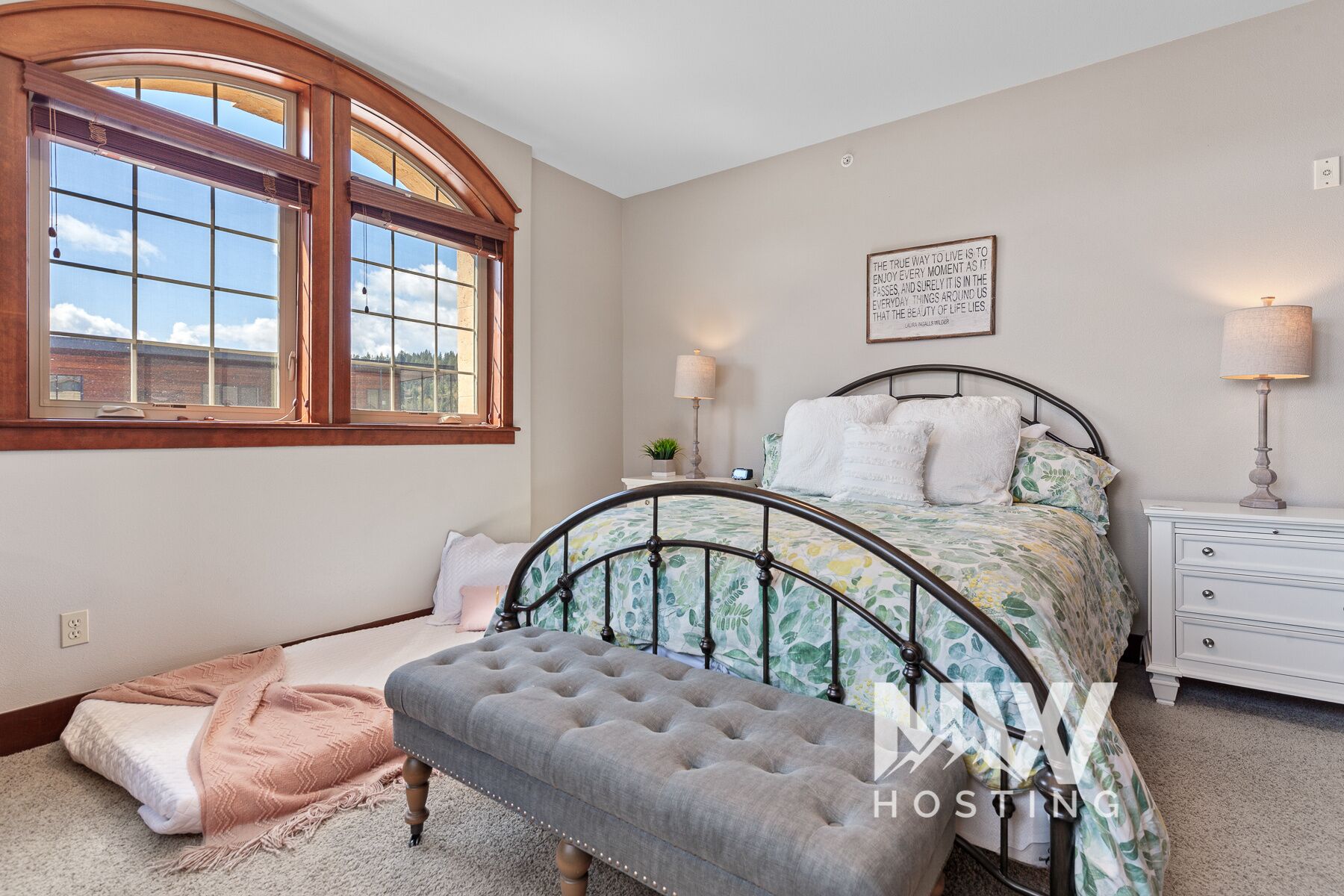 A bedroom with a large arched window, a bed with floral bedding, a bedside table with a lamp, and a small plant on the nightstand.