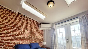 Un dormitorio con pared de ladrillo, cama con ropa de cama azul, ventana con cortinas y unidad de aire acondicionado.