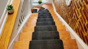 Una escalera de madera con alfombras corridas negras, plantas en macetas en el descansillo, una ventana con mosquitero y un muro de ladrillo con barandilla metálica.