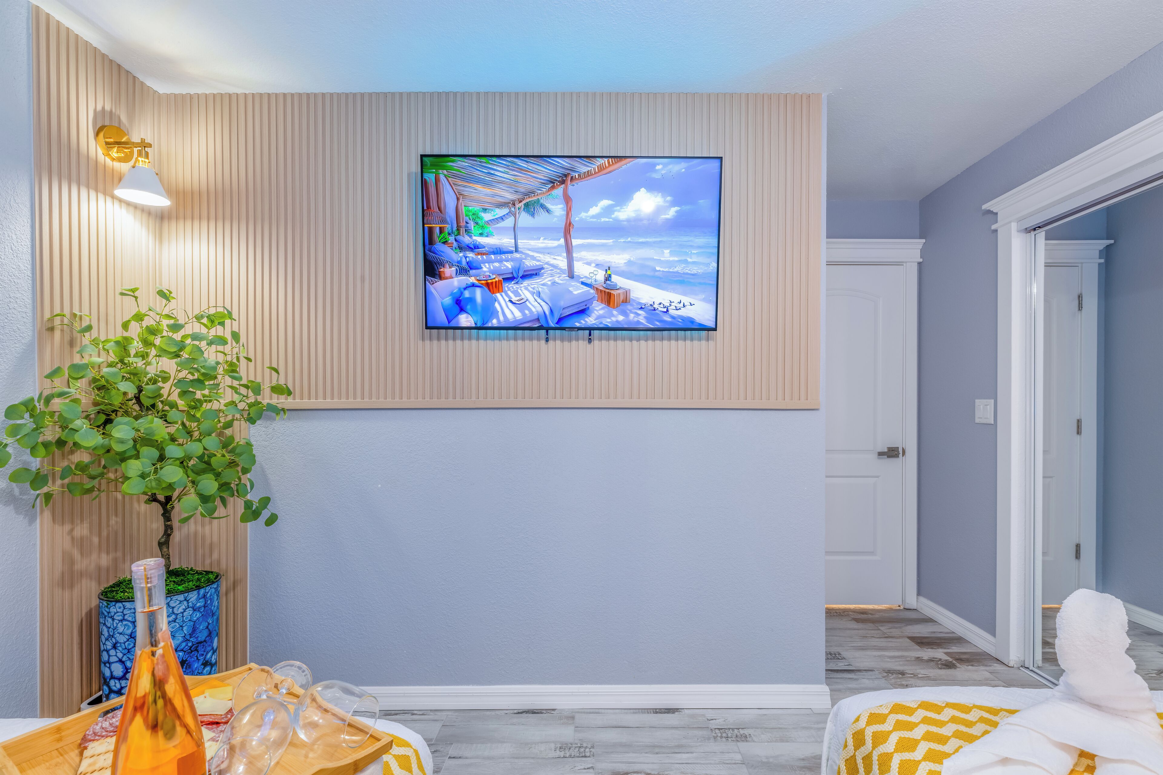 A room with a TV mounted on the wall displaying a beach scene, a potted plant, a bottle, and a tray with glasses and snacks.